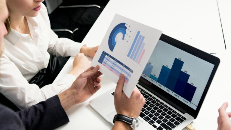 A man and a woman sit at a conference table reviewing reports on paper and on a laptop.